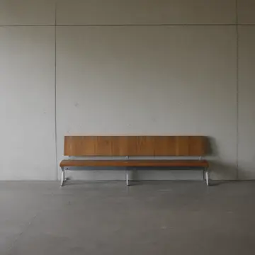 A row of empty wooden benches in Waiting room near public transit entrance. The bench slats are slowly being polished and re-aligned by an unseen mechanism. Scuff marks on the tile...
