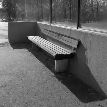 A bench seat in Bus stop shelter corner. The late afternoon cleaning crew is finishing up. Yellowed timetable poster