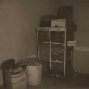 Cardboard file boxes in Office supply closet. A clerk is organizing old records, stacking them too neatly. Dust motes suspended in weak light