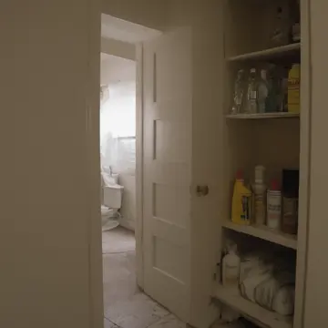 A stack of mismatched cleaning supplies in A narrow utility closet. The room is being prepared for an anticipated, but unseen, person. Yellow-taped utility shelves
